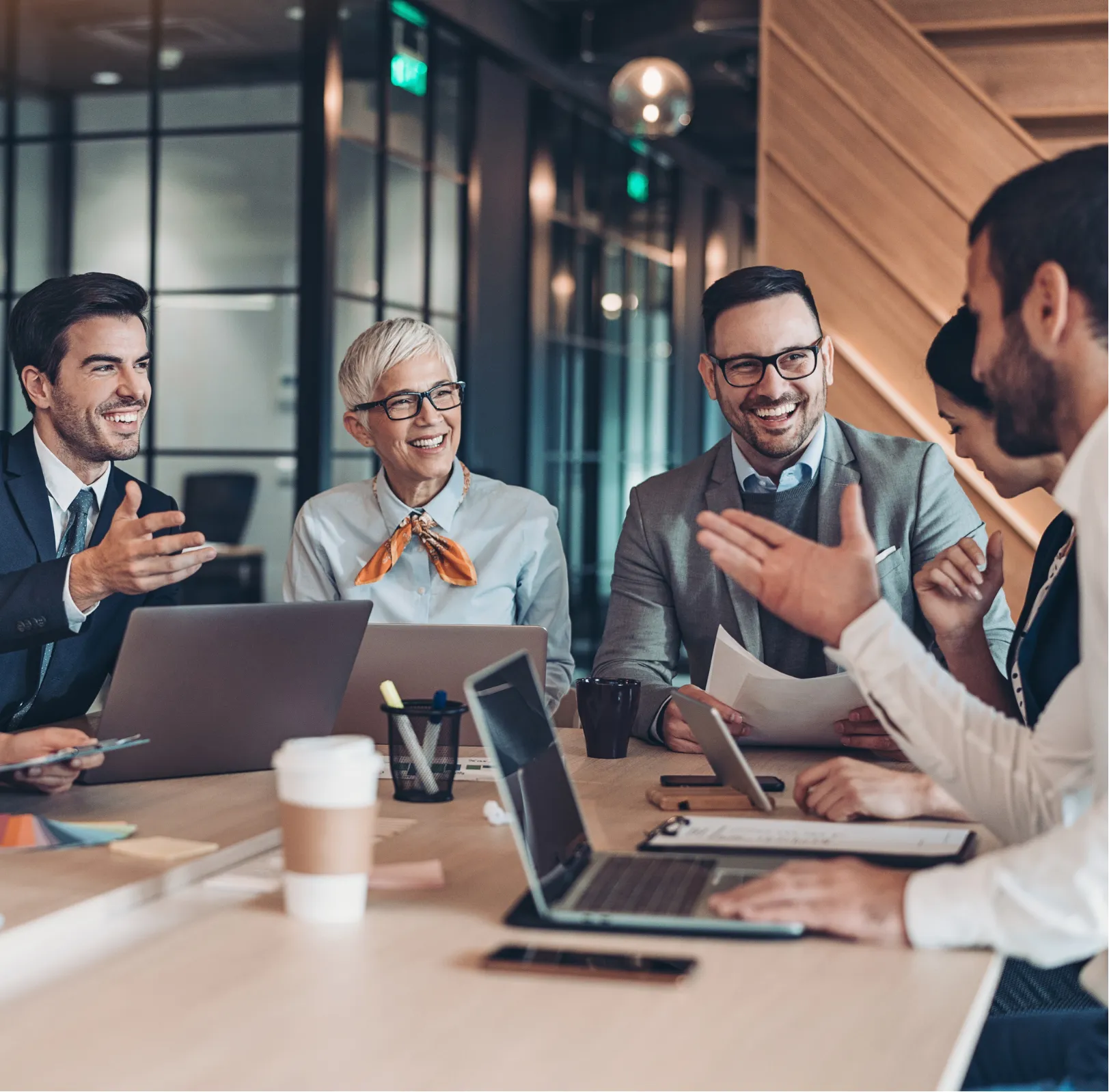 Five people in business attire smiling and talking around a meeting table with laptops and papers.