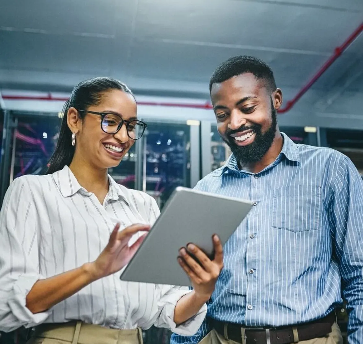 Two professionals smiling and looking at a tablet in a modern office or server room setting.