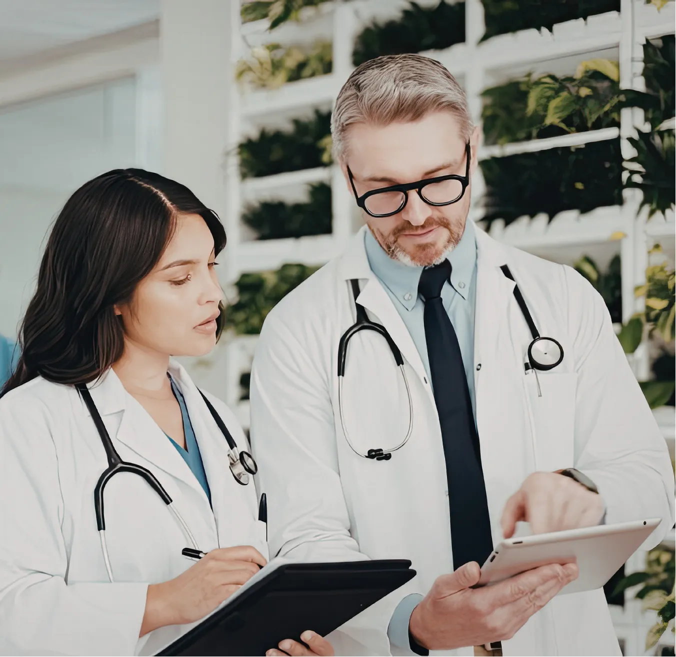 Two doctors with stethoscopes review notes together on a tablet and clipboard in a modern clinic.