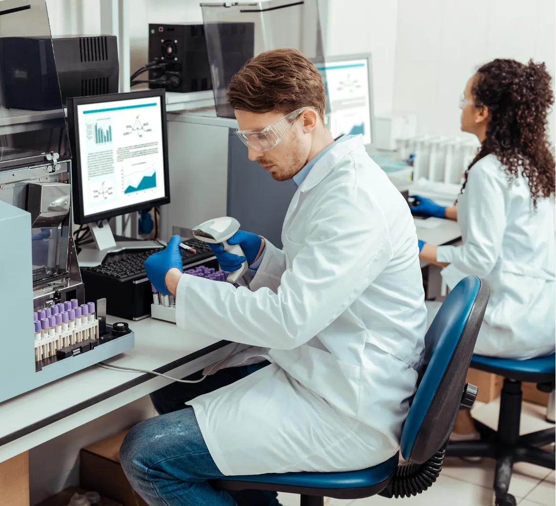 Two scientists in lab coats work at computers with test tubes and lab equipment in a laboratory setting.