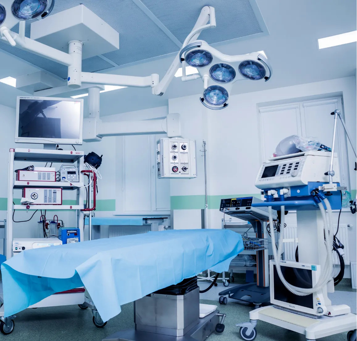 Empty operating room with surgical table, medical equipment, monitors, and overhead surgical lights.