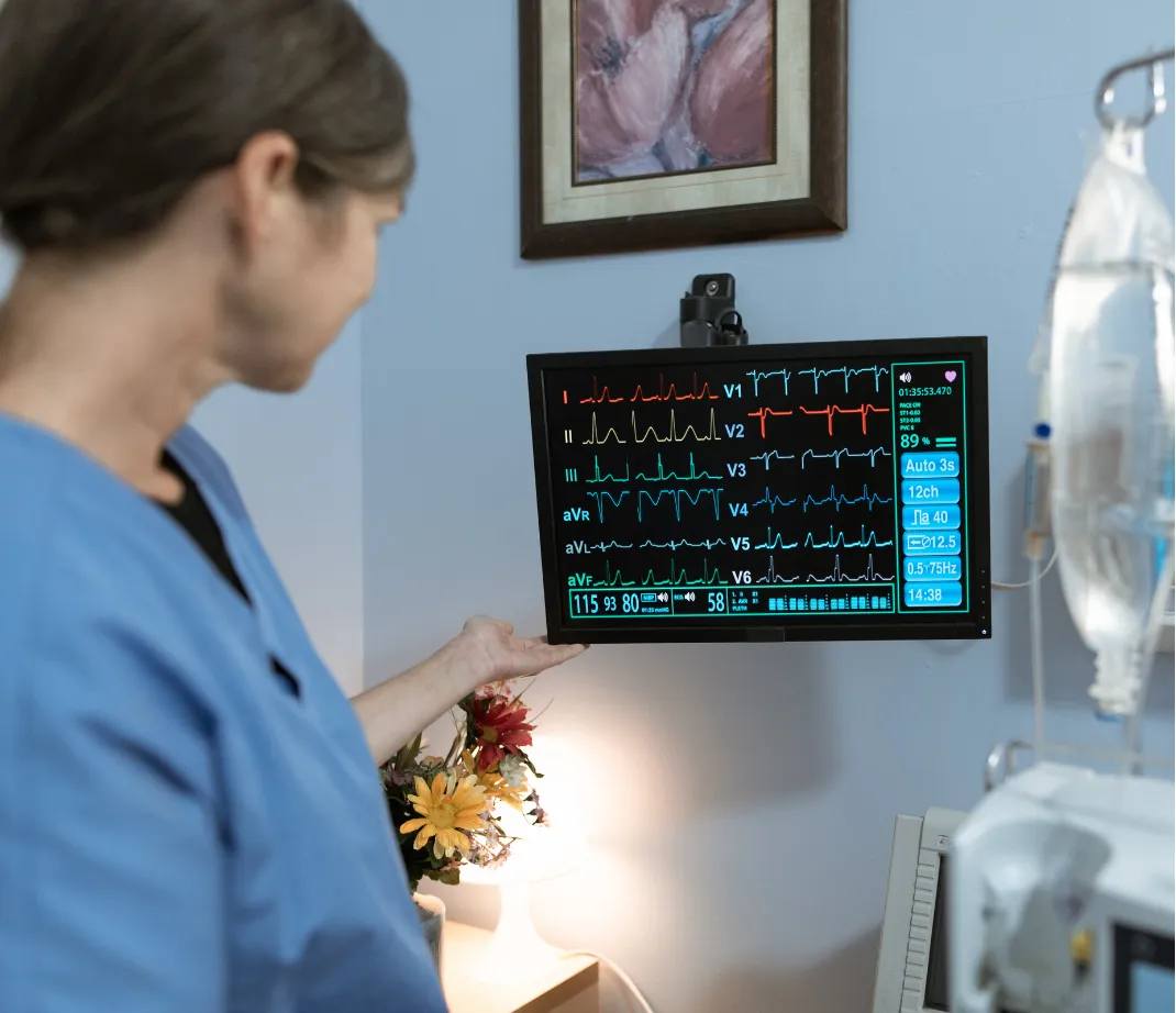 Nurse in blue scrubs looking at a heart rate monitor screen in a medical room.